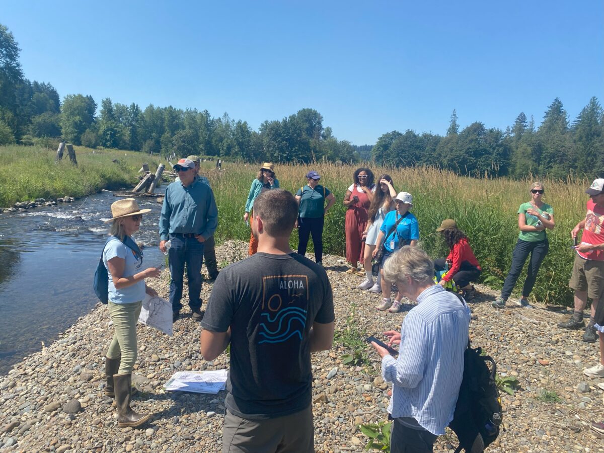 Kristin Williamson describe the processes of stream channel rebuilding while standing along side a new side channel to South Prairie Creek. She is surrounded by a group of about 15 people all standing on a gravel bar next to the creek. There are tall green plants behind the group and the sky is a bright blue.